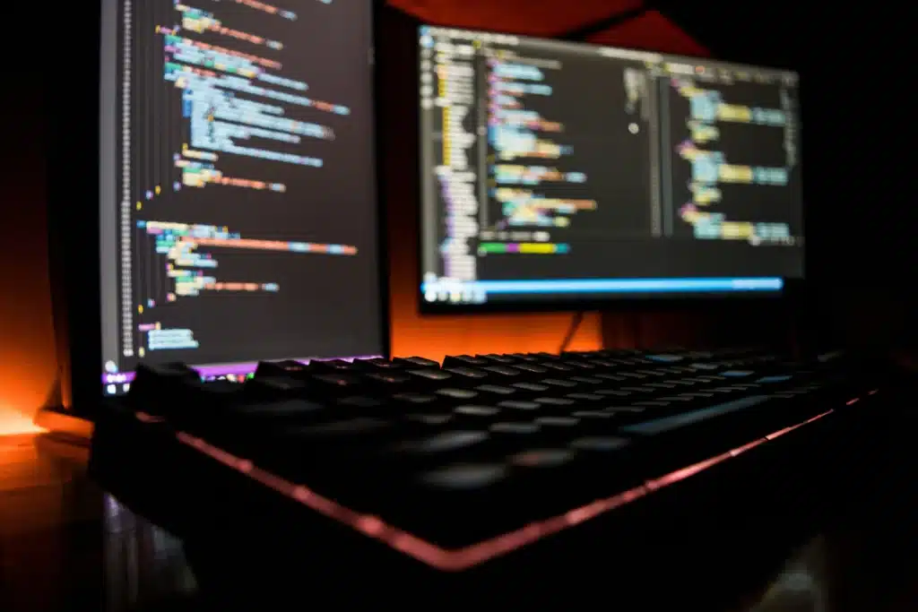 Strony internetowe i sklepy online dla firm w Polsce - Kod źródłowy na dwóch monitorach w ciemnym pomieszczeniu – stanowisko programisty z podświetlaną klawiaturą.